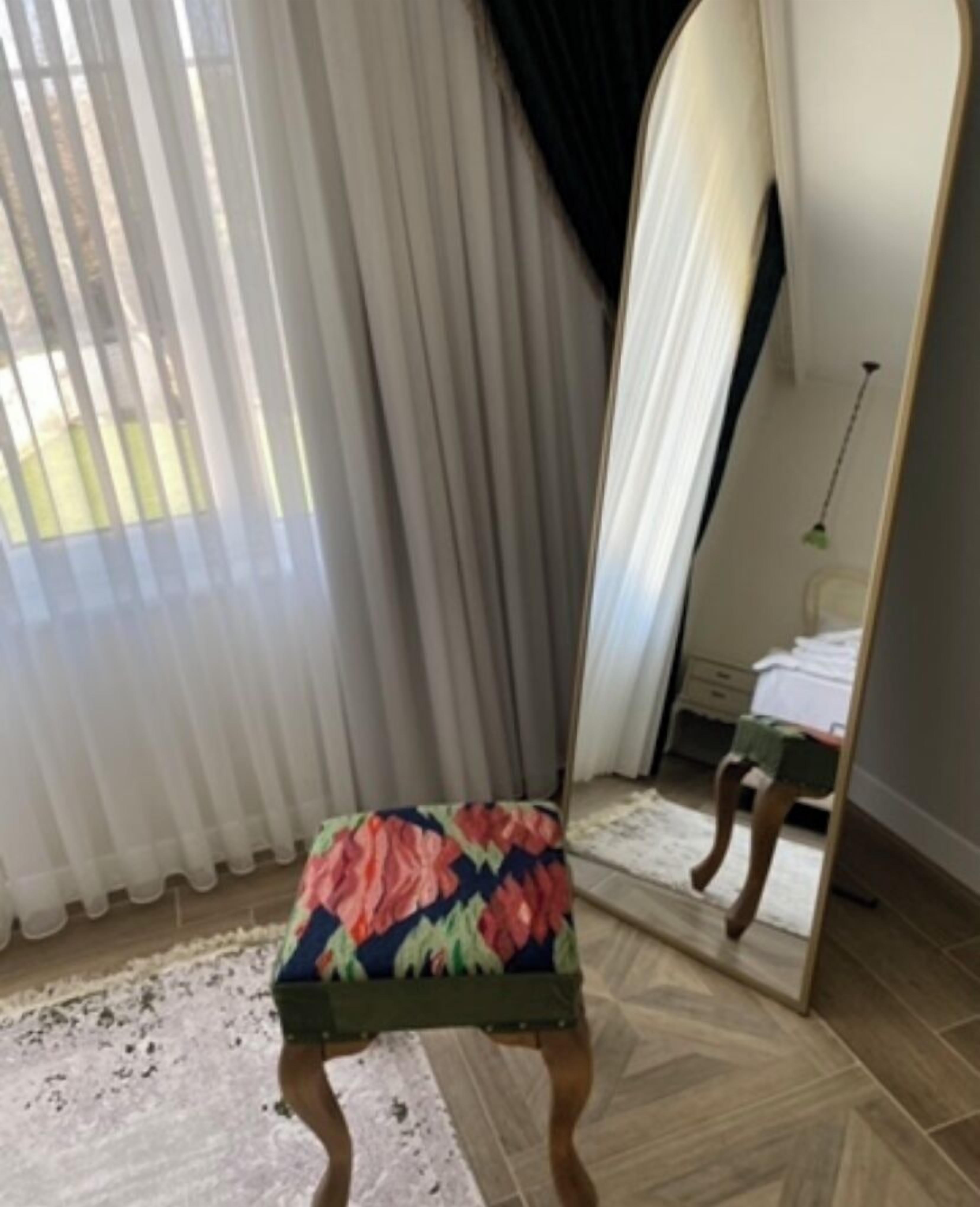 A room with a chair in front of a window with sheer curtains, a mirror reflecting part of the room, and a wooden floor.