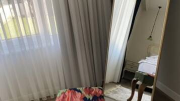 A room with a chair in front of a window with sheer curtains, a mirror reflecting part of the room, and a wooden floor.