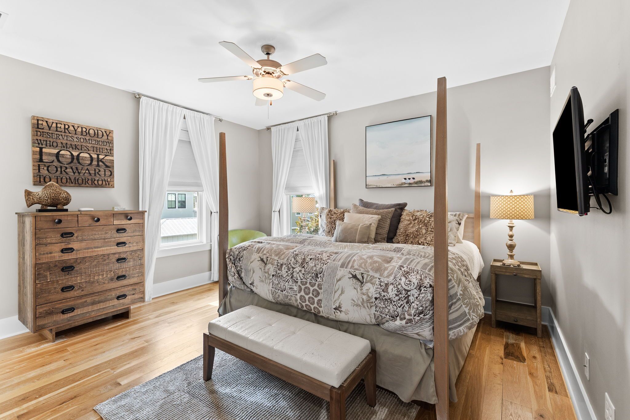 A bedroom with a four-poster bed, a wooden dresser, a bench, a ceiling fan, a TV, and a window with curtains.