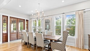 A dining room with a wooden table and six chairs, large windows with curtains, and a chandelier.