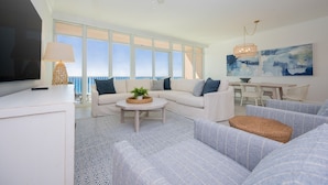A modern living room with a white sofa, a coffee table, and a TV. There is a large window with a view of the ocean. - Phoenix Gulf Towers 906 2 Bedroom Condo (Orange Beach)