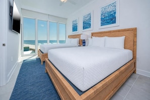 A bedroom with two beds, a large window with a beach view, and artwork on the wall.