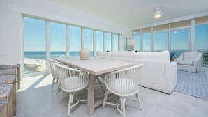 A spacious living area with a large window overlooking the beach, a white sofa, a dining table with chairs, and a ceiling fan. - Phoenix Gulf Towers 908 4 Bedroom Condo (Orange Beach)