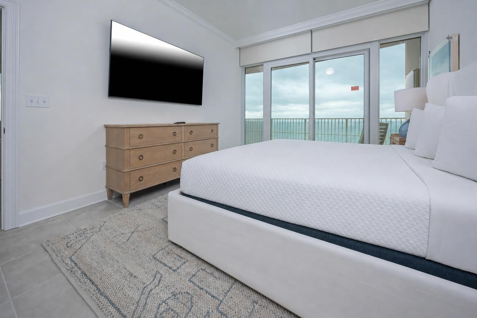 A bedroom with a large bed, a dresser, a flat-screen TV, and a balcony with a view.