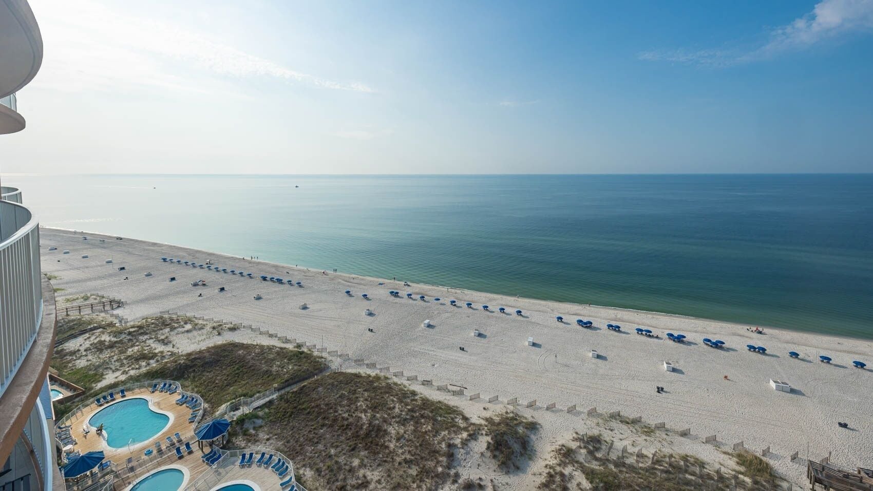 A beach with sun loungers and a pool area, viewed from a balcony.