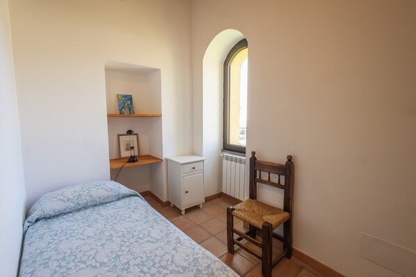Un dormitorio con una cama, una silla, un pequeño tocador blanco, un estante con un cuadro y una ventana con arco.