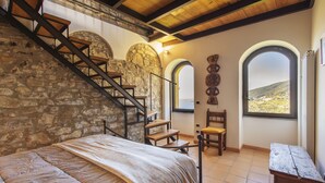 Un dormitorio con pared de piedra, escalera de madera y vista al campo a través de ventanales abovedados.