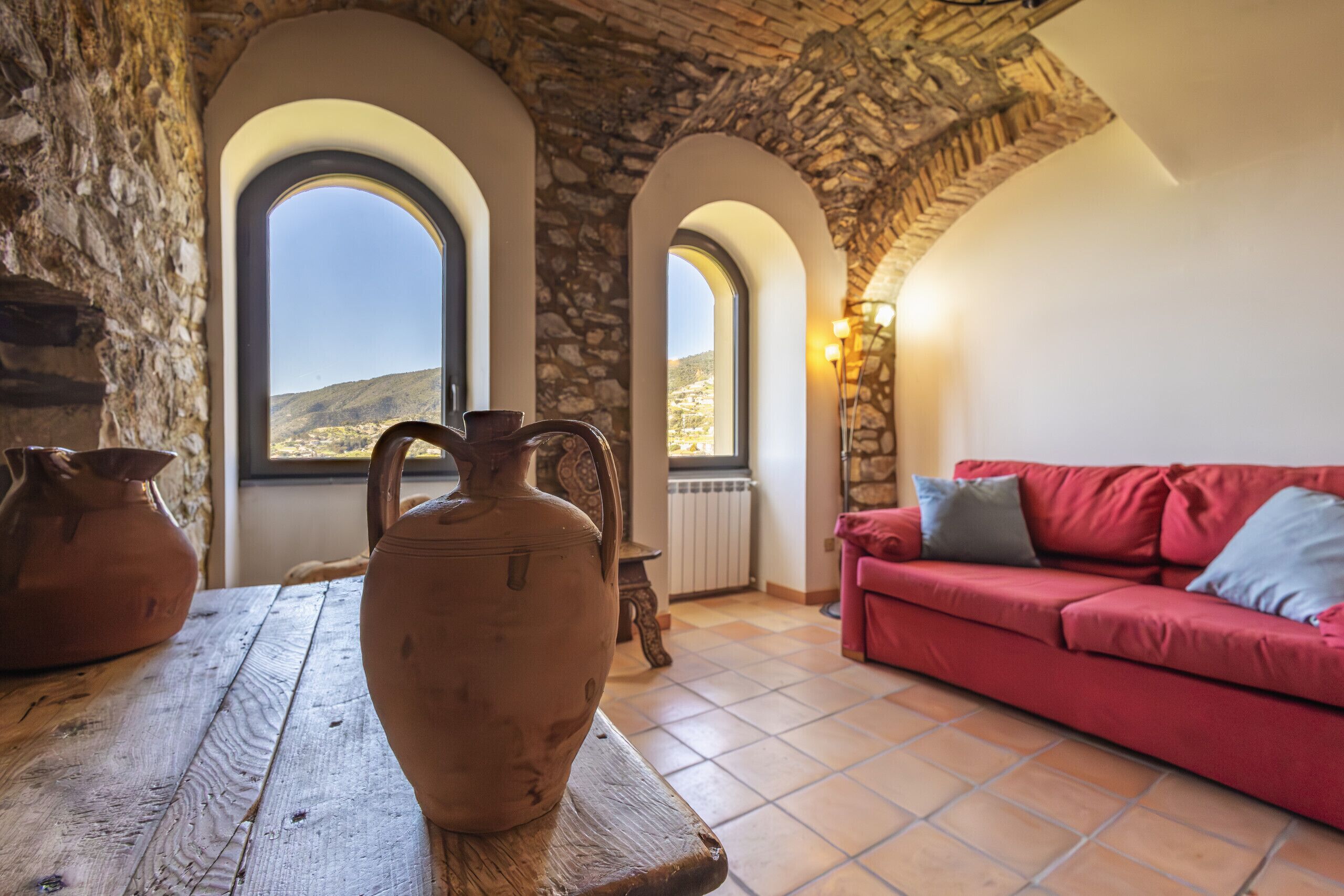A room with a stone wall, a red sofa, a wooden table with a clay pot, and arched windows with a view of the countryside.