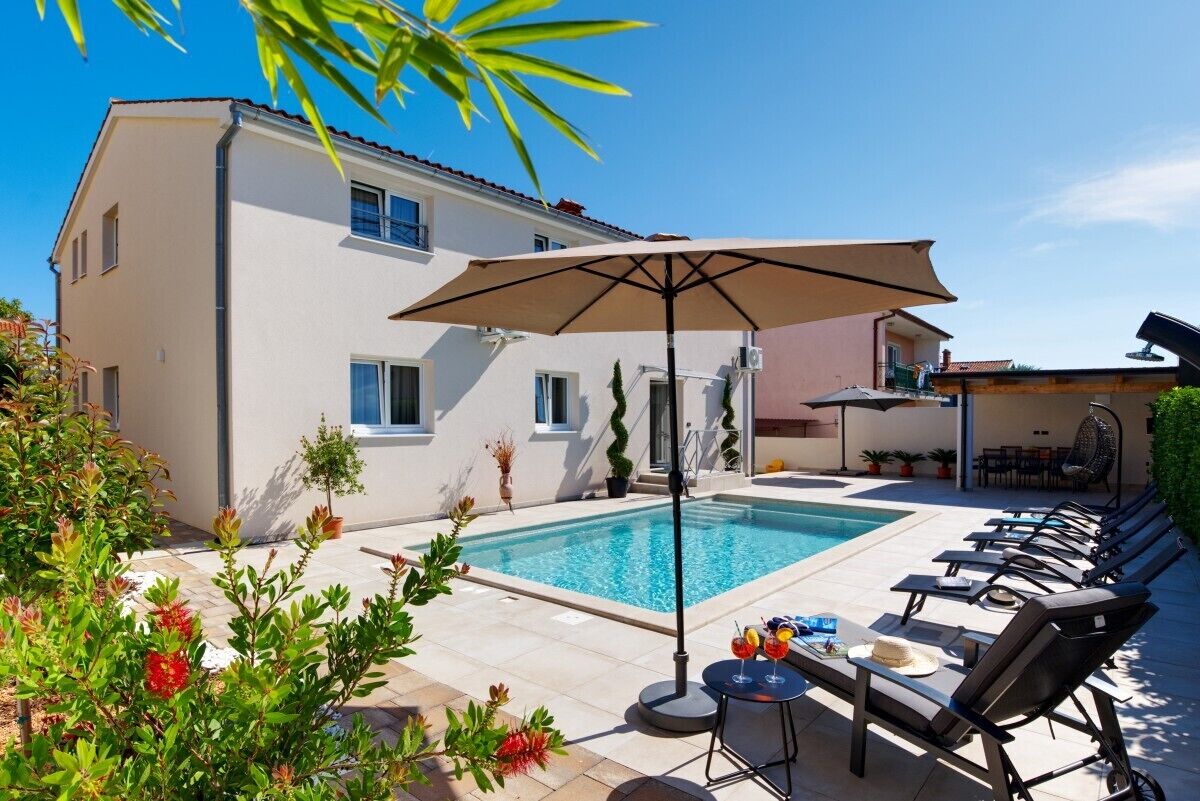 A swimming pool with lounge chairs and a table, surrounded by a building with a tiled patio.