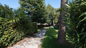 Un chemin pavé, bordé d’une végétation luxuriante et de grands arbres, mène à un bâtiment à la façade blanche.
