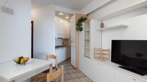 Une cuisine compacte avec des meubles blancs, une table à manger avec des chaises, un panier de fruits, un téléviseur et une petite étagère avec une plante.