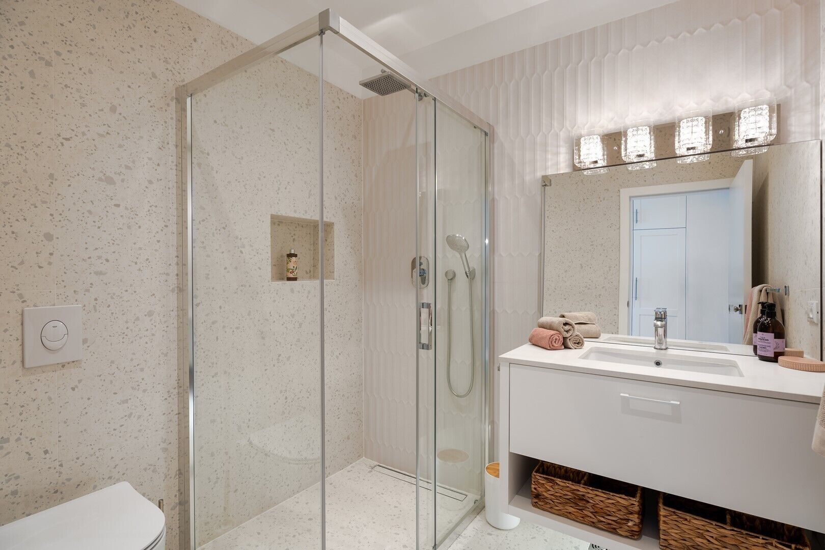 A modern bathroom with a glass-enclosed shower, a white sink, and a large mirror with lights above it.