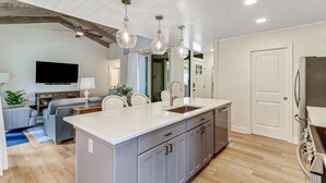 A modern kitchen with a central island, grey cabinets, and stainless steel appliances. There is a dining area with a grey sofa and a TV mounted on the wall.