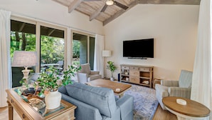 A spacious living room with a ceiling fan, a flat-screen TV mounted on the wall, and a large window providing a view of the outdoors.