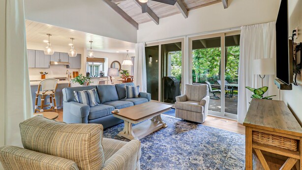 A spacious living area with a blue sofa, a wooden coffee table, and wicker chairs. There is a sliding glass door leading to an outdoor patio.
