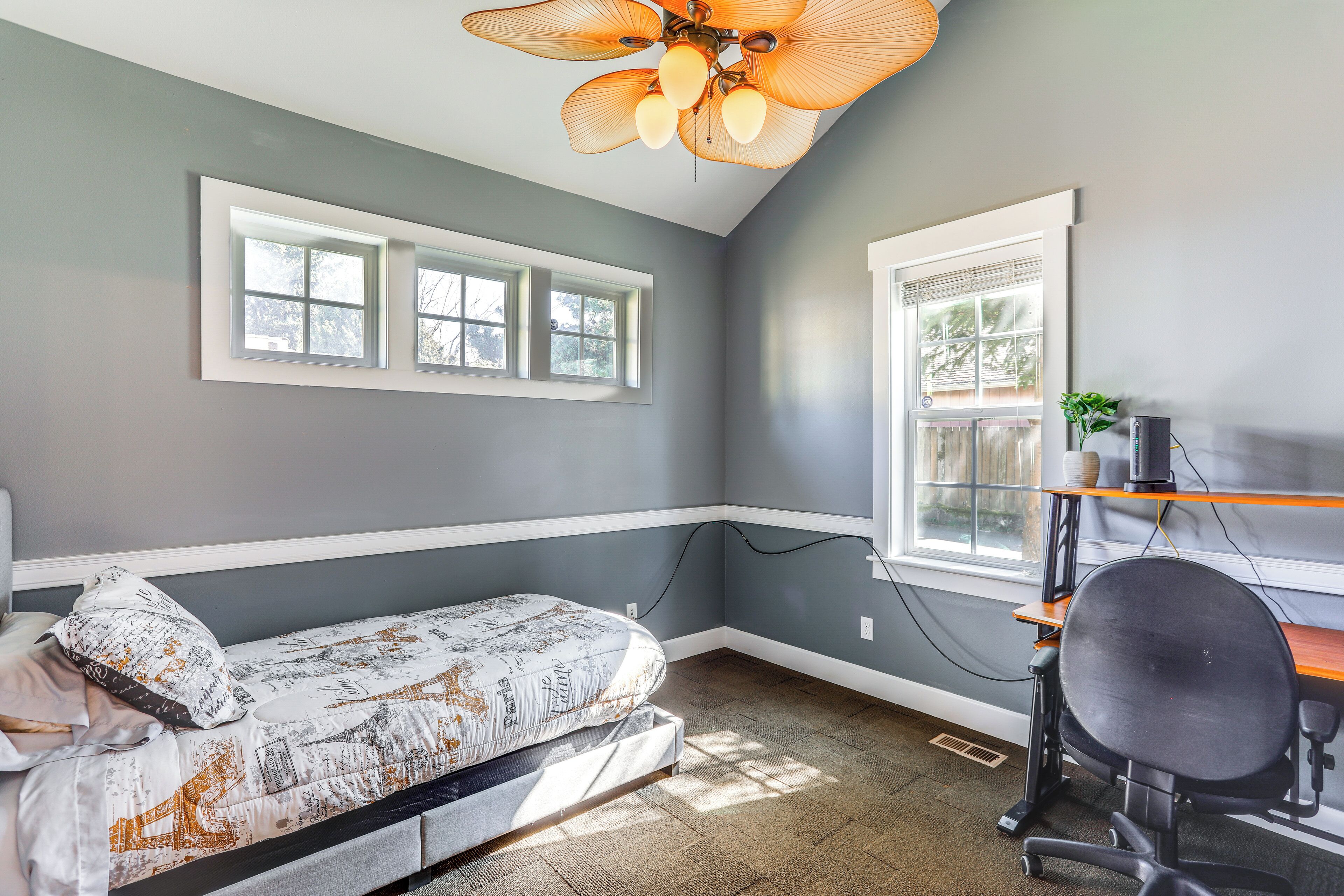 A bedroom with a bed, a desk, and a chair. There are two windows and a ceiling fan.