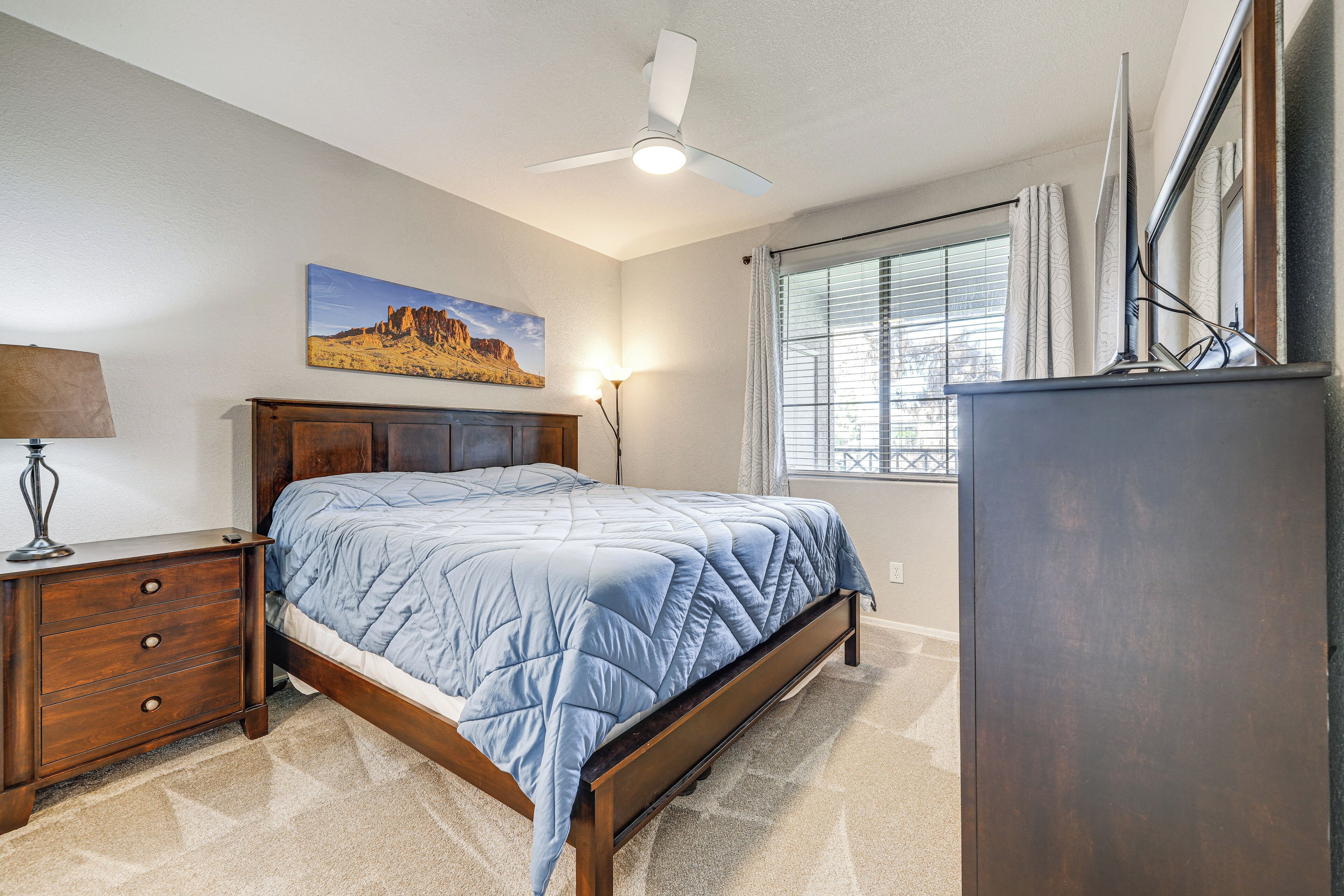 A bedroom with a bed, a nightstand, a dresser, a ceiling fan, a window with curtains, and a painting on the wall.