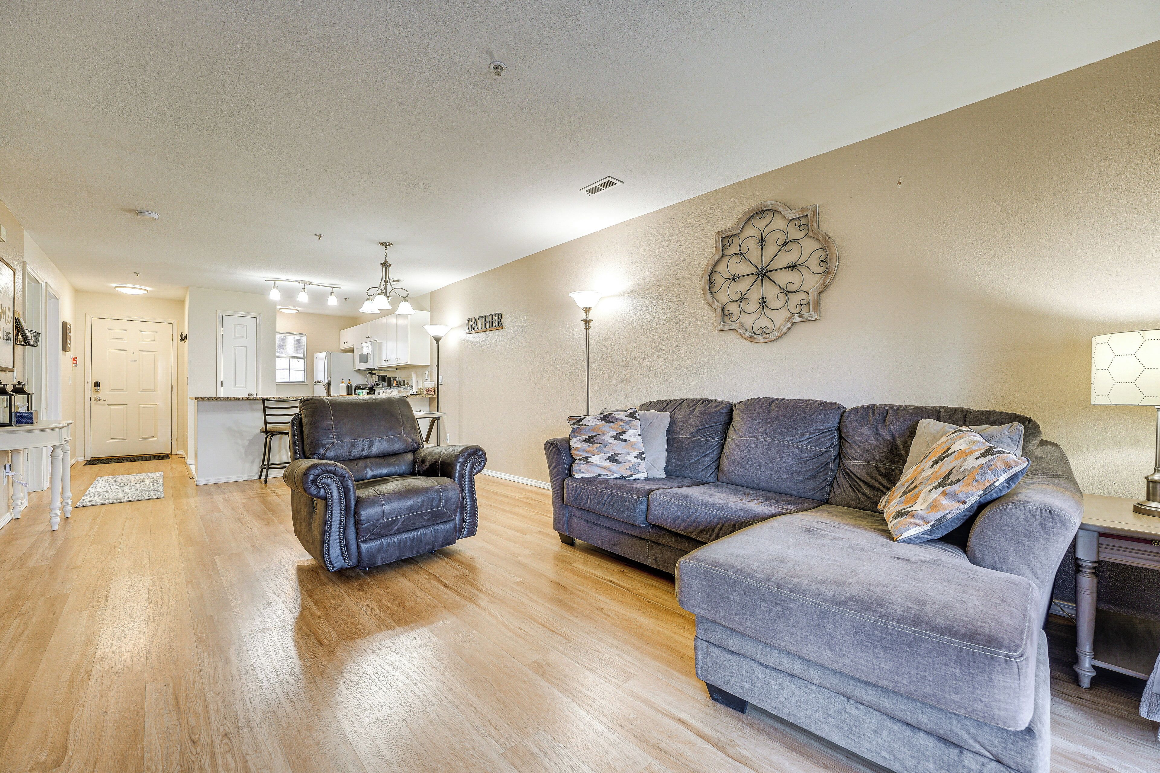 A spacious living room with a leather armchair, a grey sofa, and a matching ottoman. A kitchen is visible in the background with a white door and a ceiling light fixture.