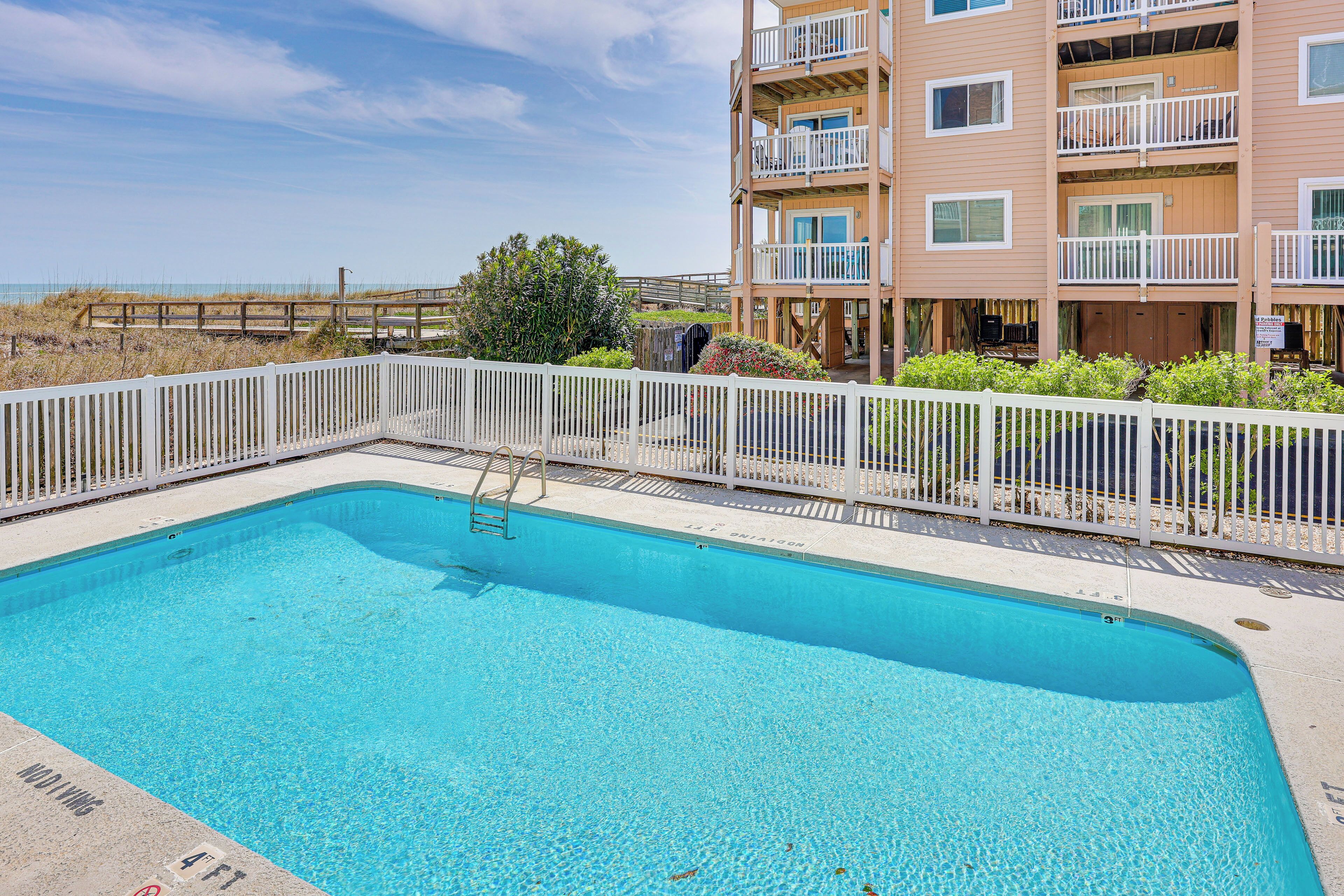Une piscine rectangulaire entourée d’une clôture blanche, avec une terrasse en bois et, au fond, un bâtiment doté de balcons.