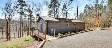 A cabin with a gravel driveway, a wooden deck, and a view of a lake surrounded by trees.