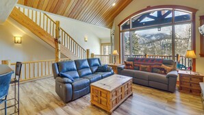 Un vaste salon doté d’un plafond en bois, d’une grande fenêtre donnant sur les montagnes, d’un escalier et de canapés en cuir.