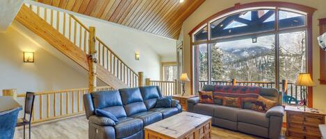 A spacious living room with a wooden ceiling, a large window offering a mountain view, a staircase, and leather sofas.