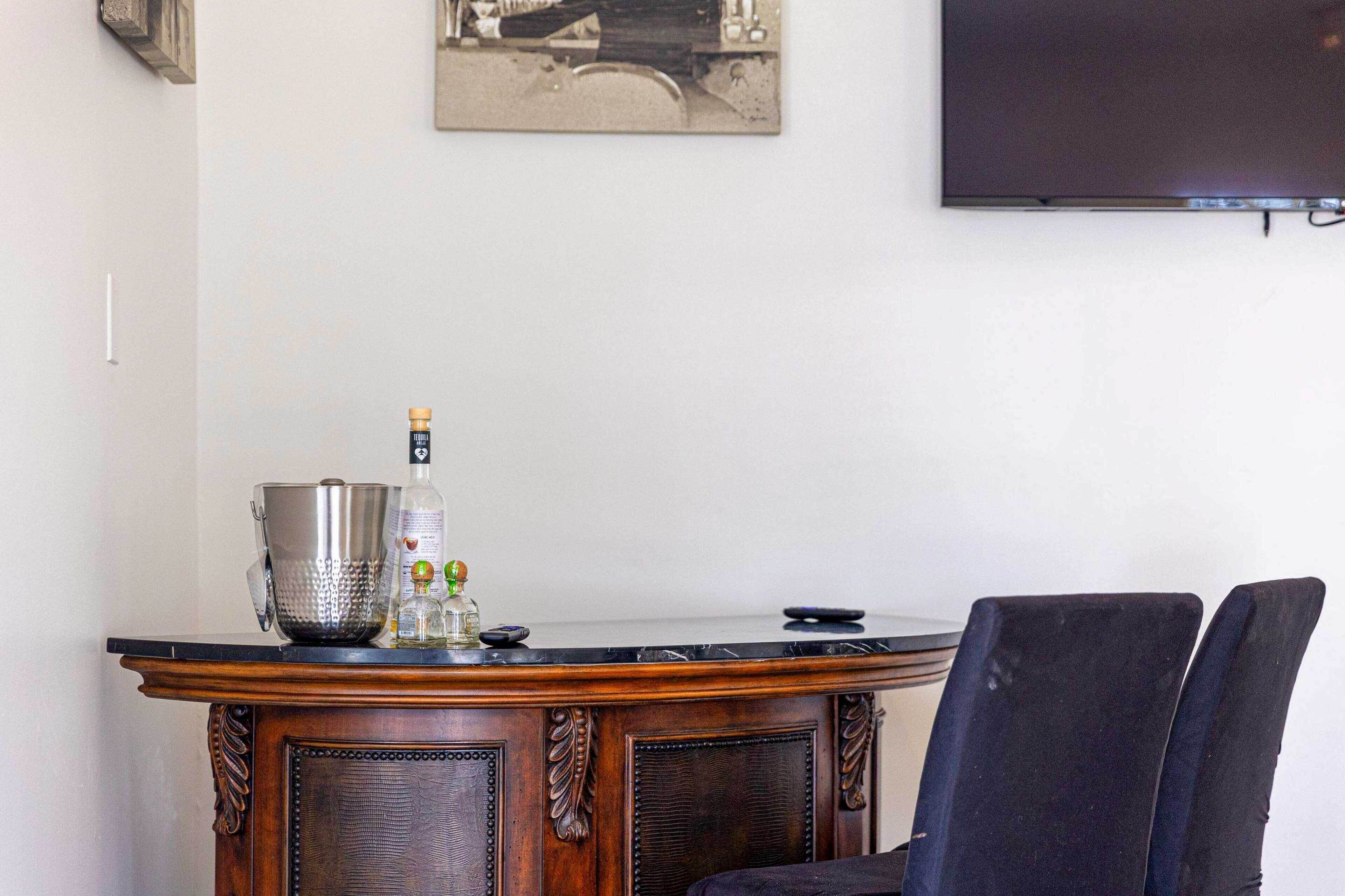 A bar area with a wooden bar counter, a large metal pitcher, a glass bottle, and a remote control. There are two dark chairs and a television mounted on the wall.