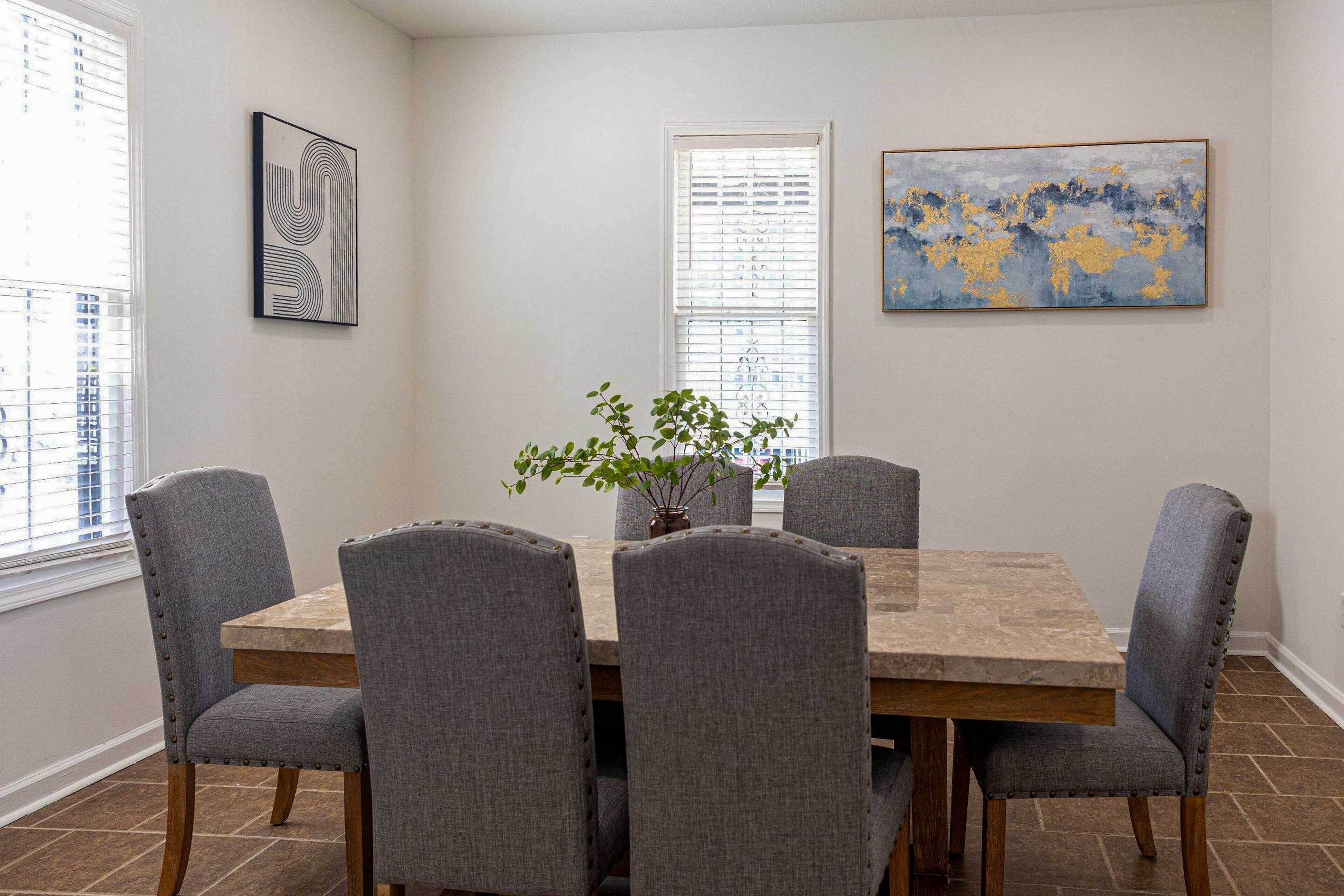 Un comedor con una mesa de madera y seis sillas grises tapizadas. Hay un florero con plantas verdes sobre la mesa.