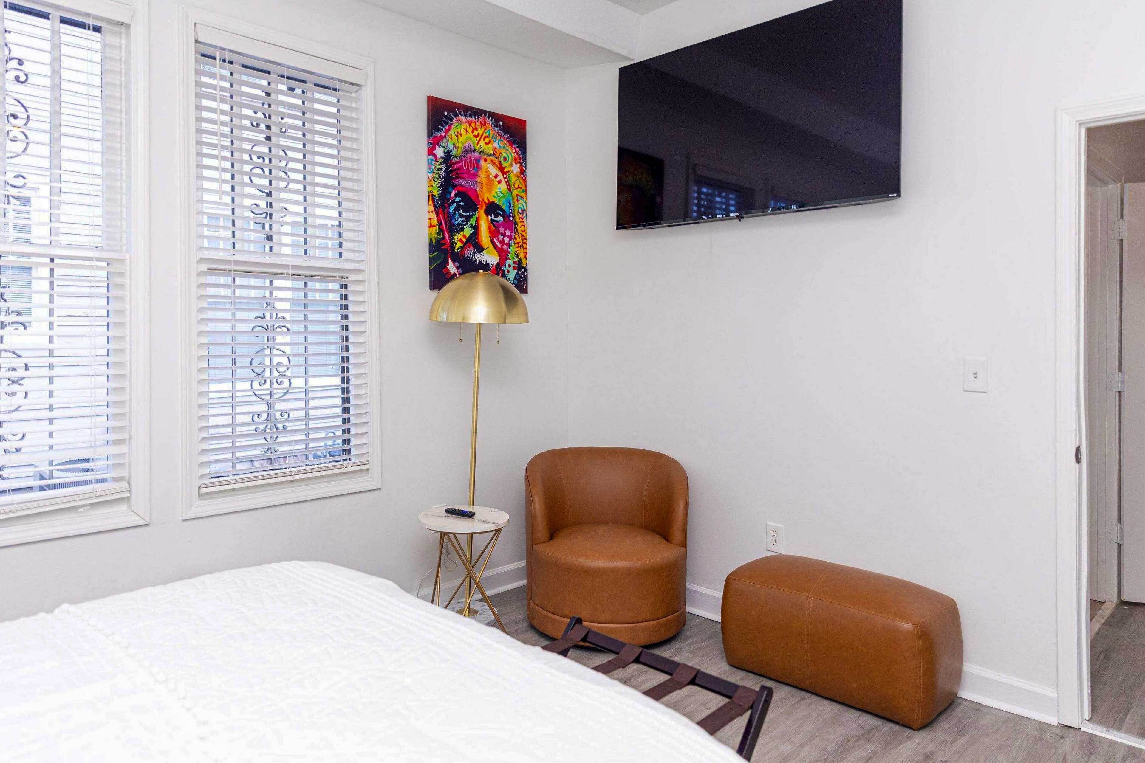 A bedroom with a bed, a brown leather chair, a small round table with a lamp, and a large TV on the wall.
