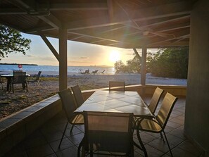 Katettu ulkoalue, jossa on istumapaikkoja ja josta avautuu näkymä rannalle ja auringonlaskuun.
