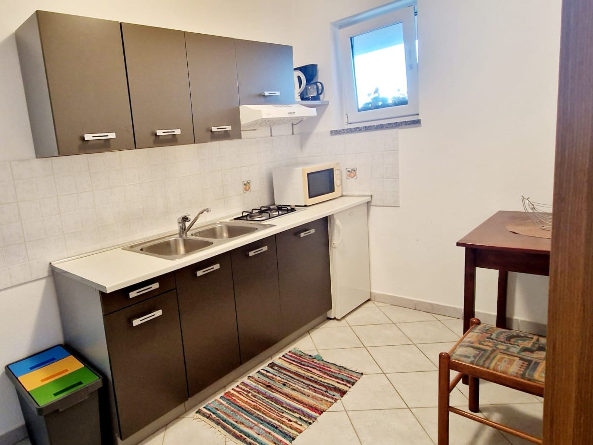 Cocina compacta con estufa a gas, dos fregaderos y microondas. Hay una pequeña ventana sobre el microondas y una silla de madera con un cojín colorido.
