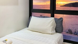 A bedroom with a large window overlooking a sunset over water, a bed with white bedding, and a painting on the wall.