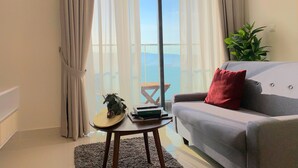 A living room with a grey sofa, a small round table with a plant and books, and a view of the sea through sheer curtains.