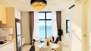 A modern hotel room with a dining area, a large window with a view, a chandelier, and a kitchenette.