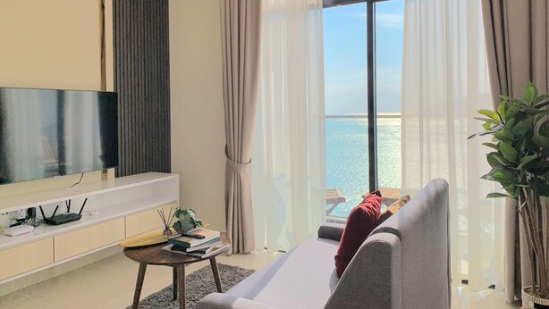 A modern living room with a large flat-screen TV mounted on the wall, a grey sofa, a small coffee table with books and a plant, and a view of the sea through large windows with sheer curtains.