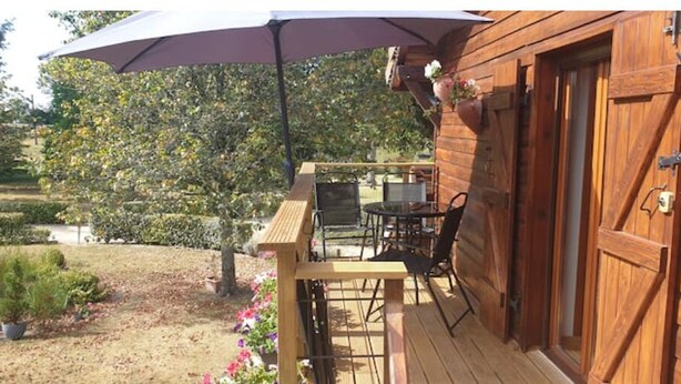 Une terrasse en bois avec une table et des chaises, un grand parasol, et un jardin agrémenté de plantes et de fleurs en pot.