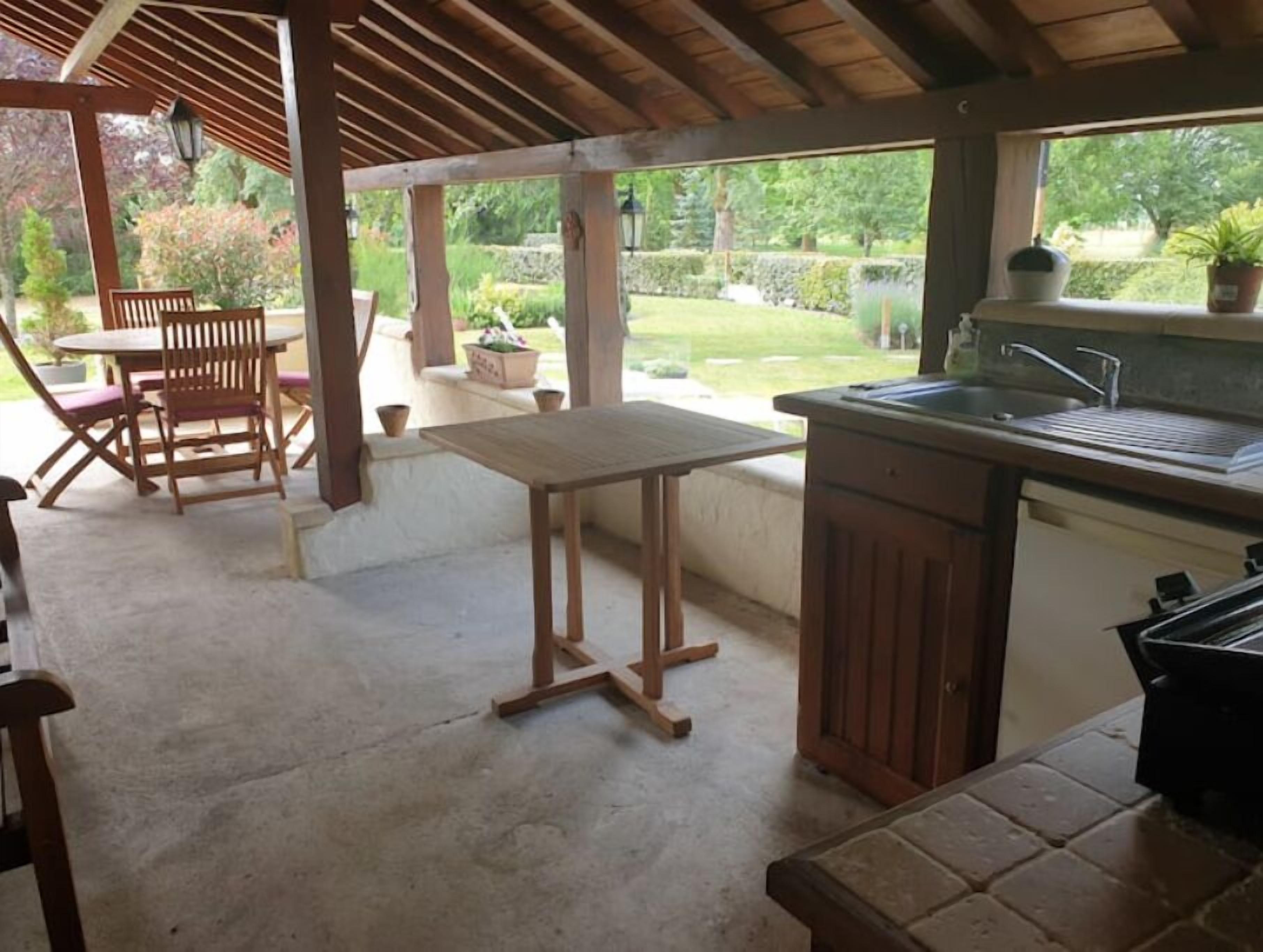 A covered patio area with a wooden table and chairs, a small kitchen with a sink and countertop, and a view of a garden.