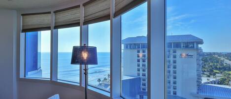 Una habitación con ventanas amplias que dan al océano, una lámpara y vista del edificio de un hotel.