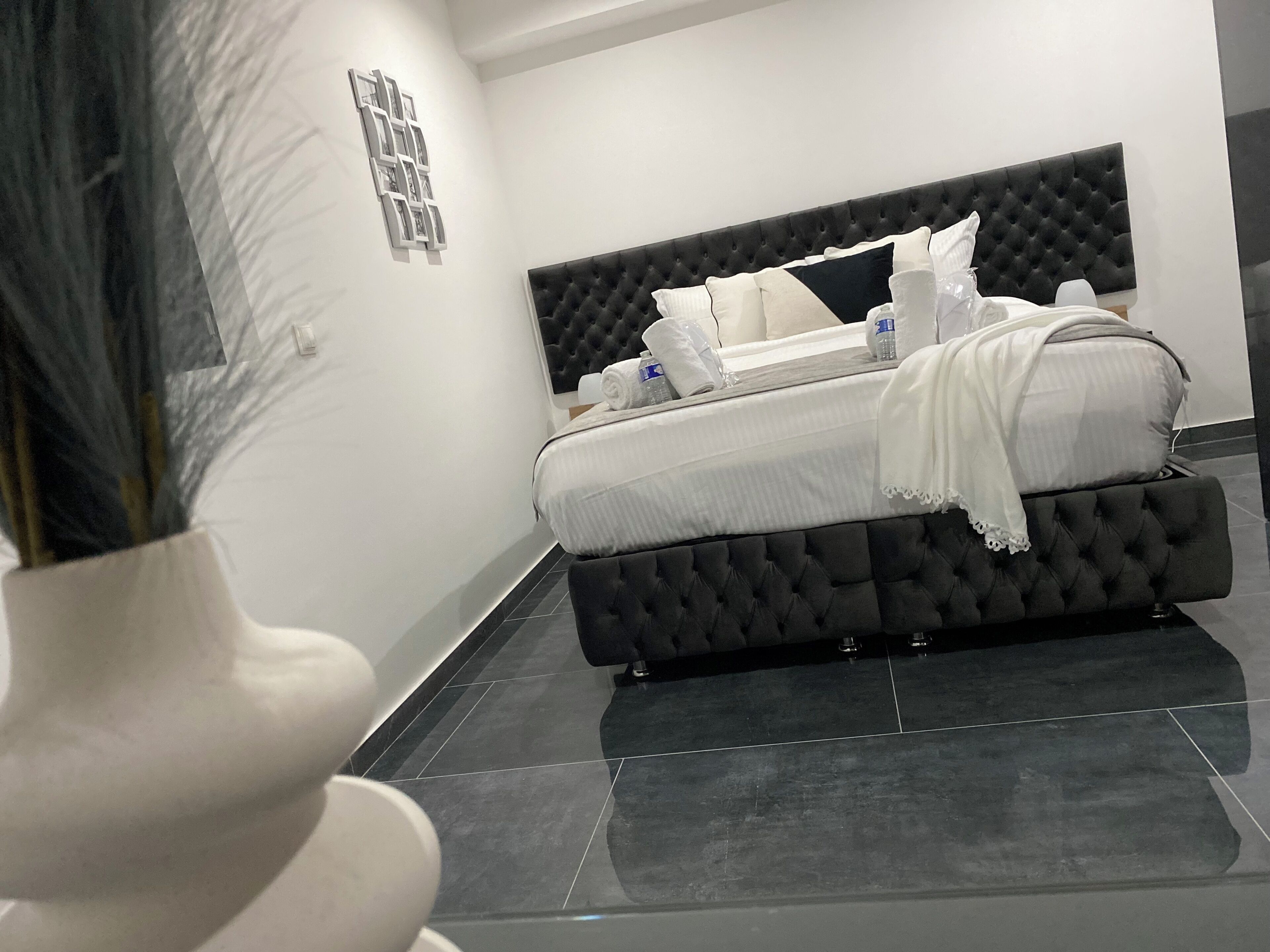 A modern bedroom with a tufted headboard, a neatly made bed with white linens, and a vase with dried plants on the left.