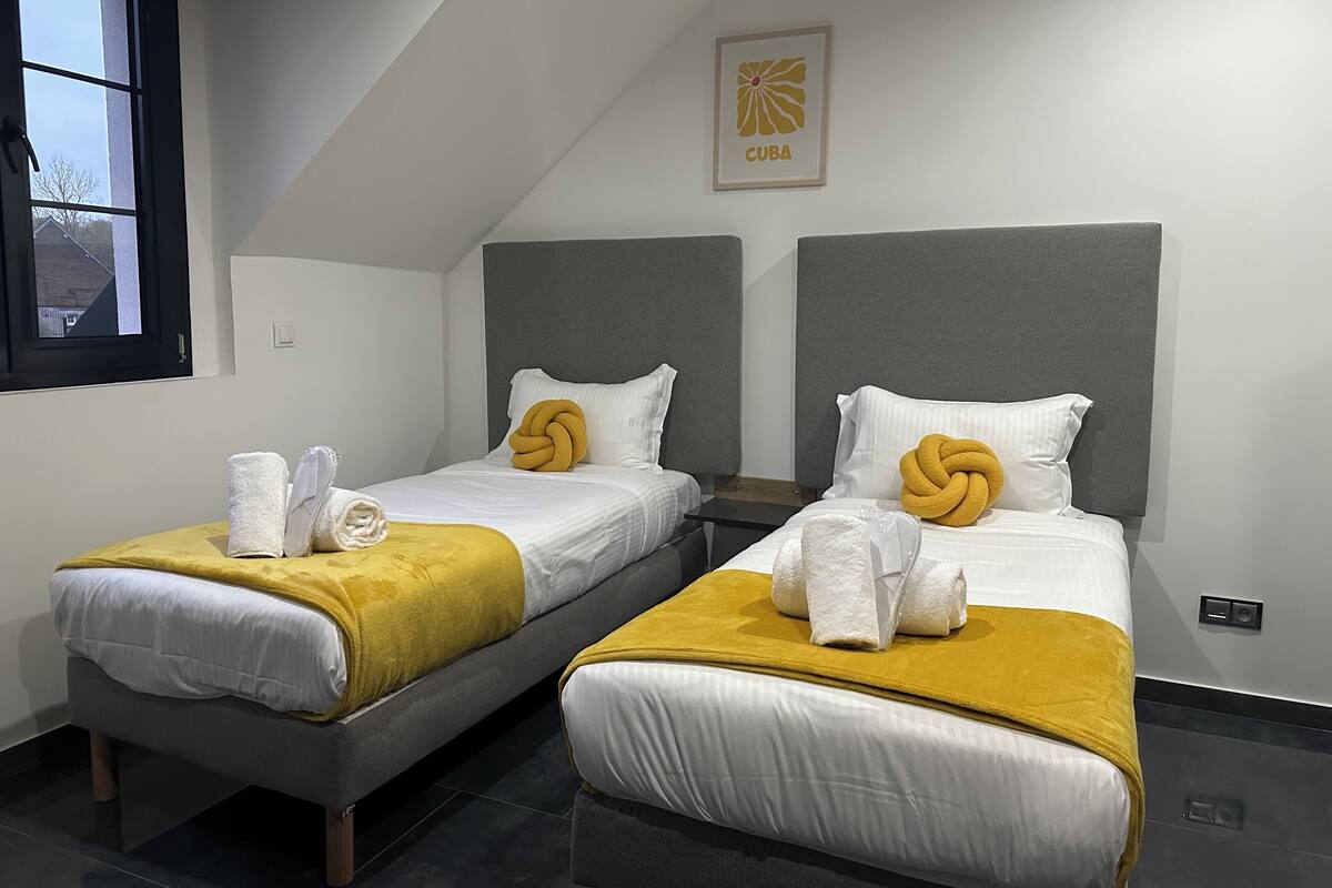 A hotel room with two beds, each with a grey headboard, white bedding, and a yellow throw blanket. There is a framed picture of a flower on the wall above the beds.