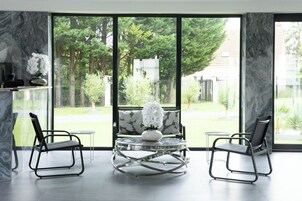 Una habitación moderna con ventanas amplias, pared de mármol y dos sillas negras alrededor de una mesa de vidrio con un jarrón blanco.