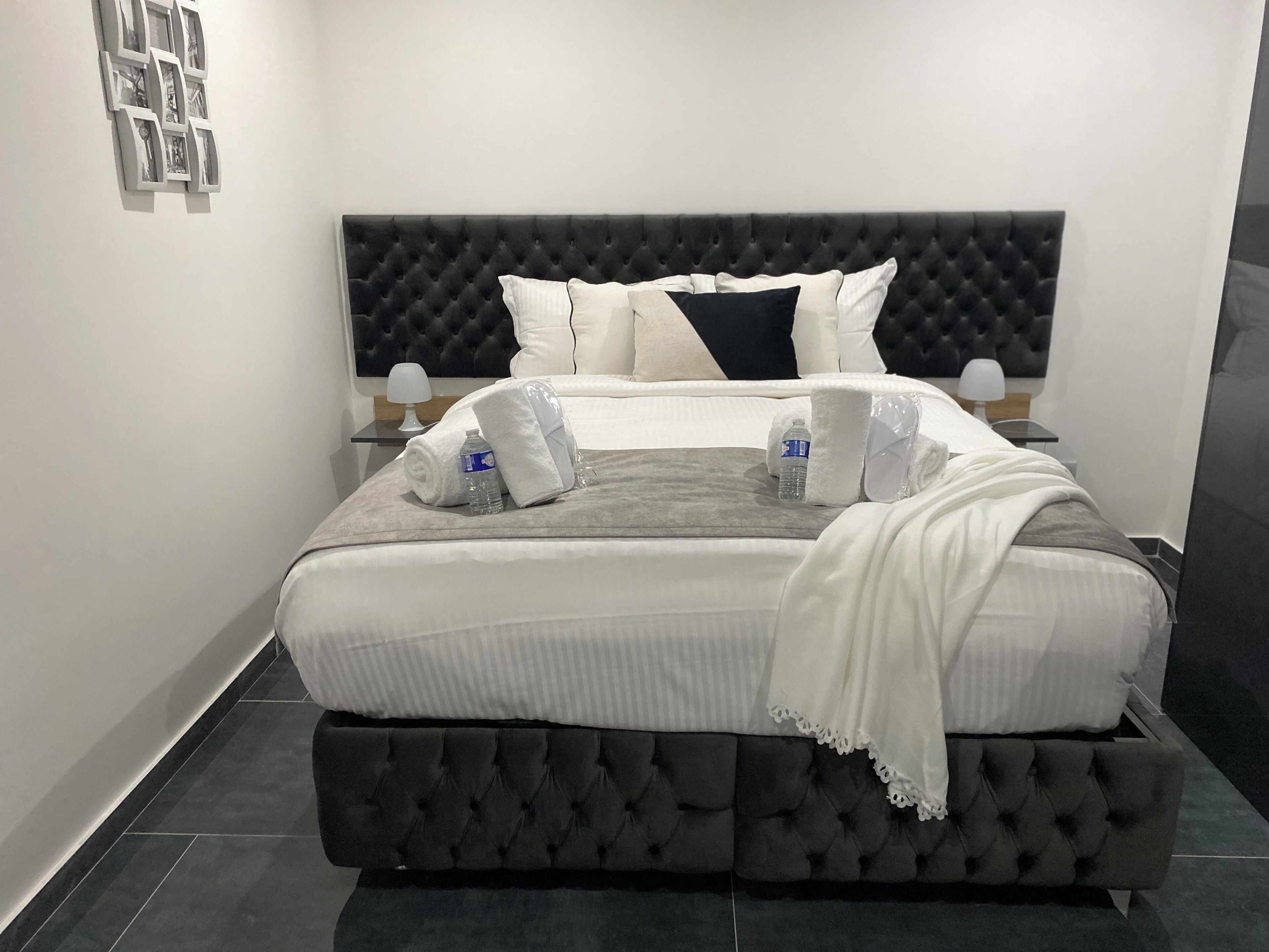 A neatly made bed with white linens, a grey throw blanket, and two towels. There are two bottles of water and a small table with a lamp beside the bed.