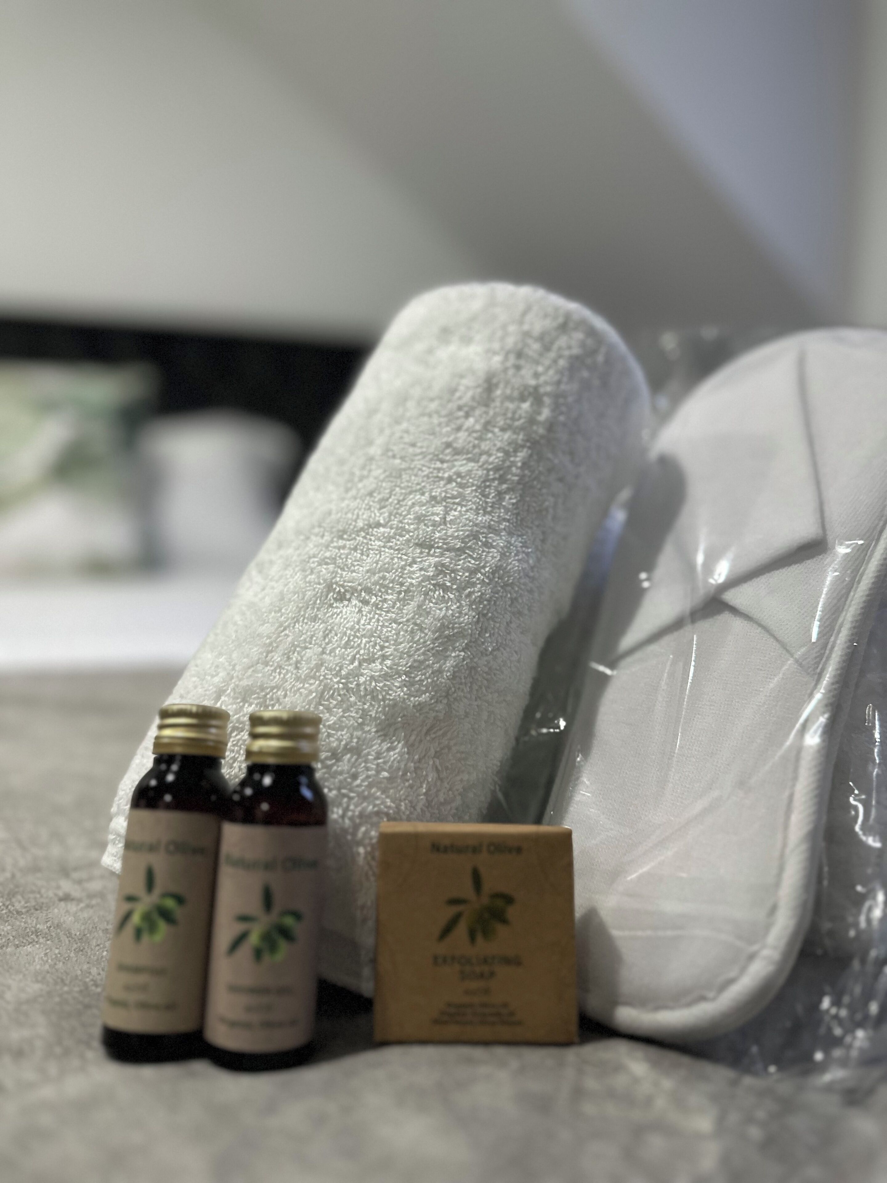 Una toalla blanca, dos botellas de aceite de oliva natural y una caja de jabón exfoliante sobre una superficie texturizada.