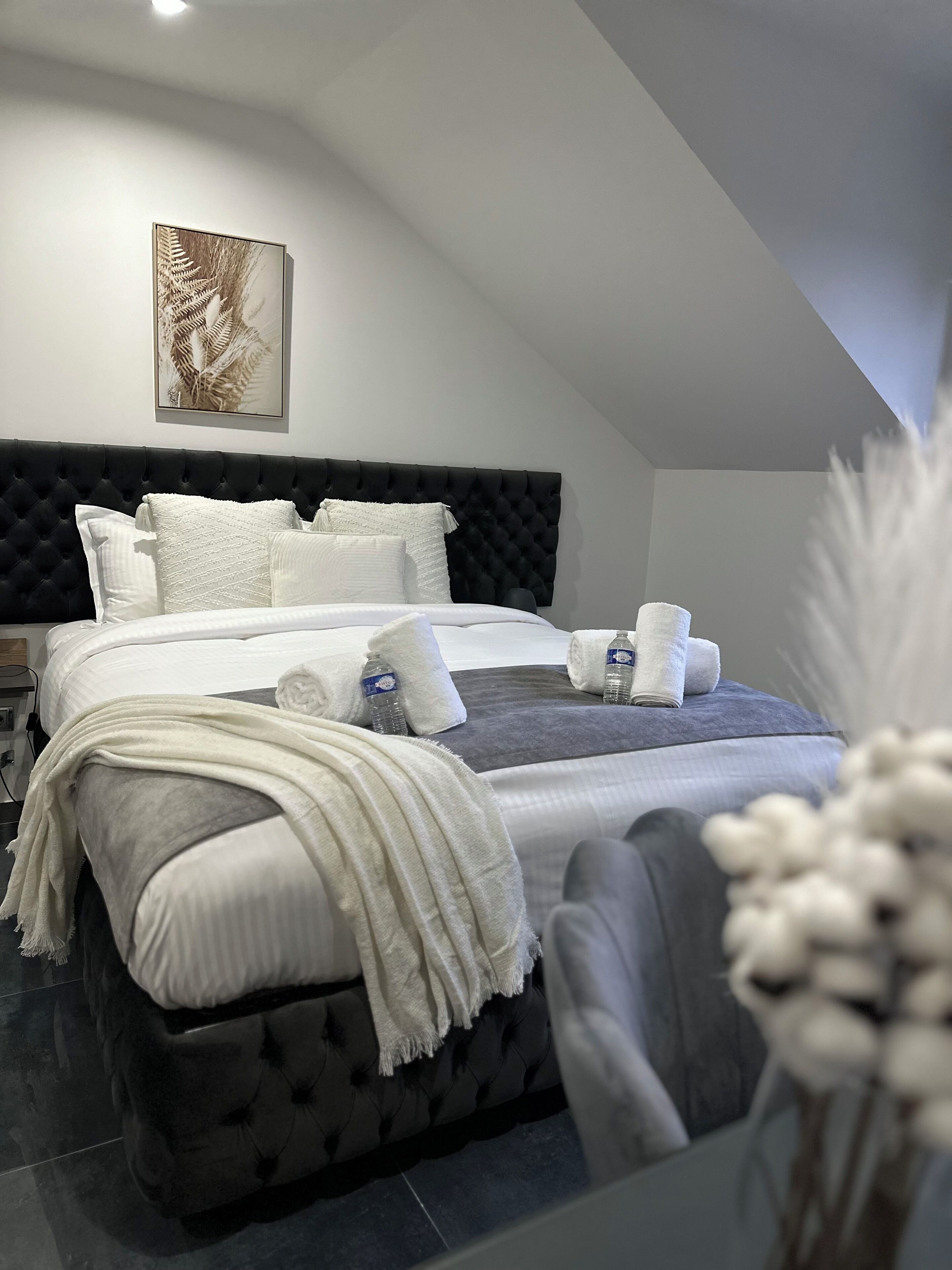 Un dormitorio moderno con cabecera capitonada, una cama grande con ropa de cama blanca, una manta gris y un jarrón blanco con flores secas.