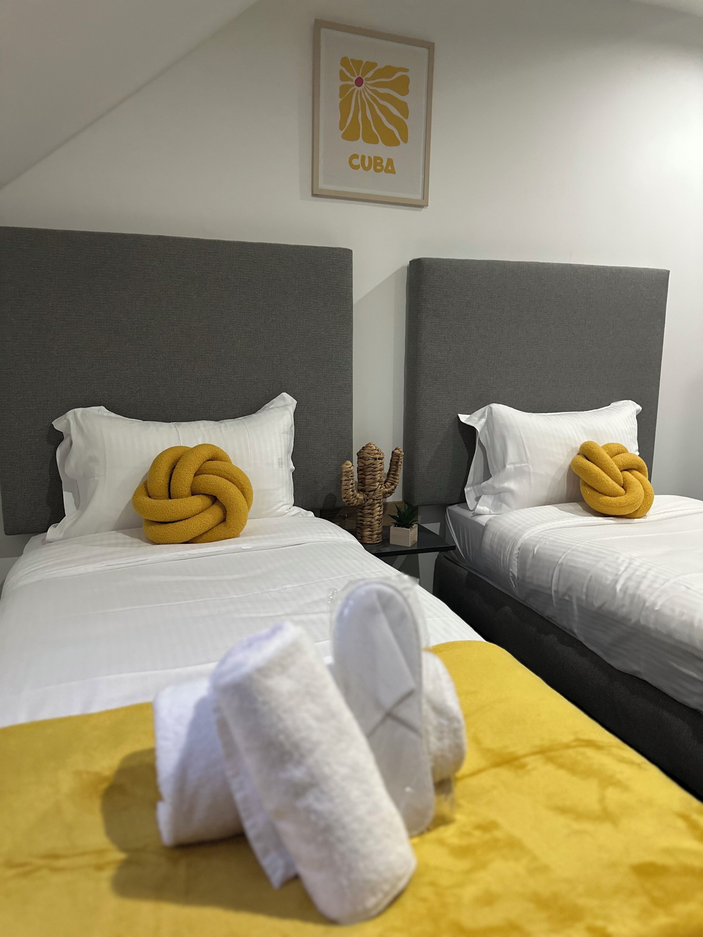 Ein Hotelzimmer mit zwei Betten, jedes mit einem einzelnen gelben Handtuch und weißen Kissen. An der Wand hängt ein gerahmtes Bild einer Blume, mit dem Schriftzug 'CUBA'.