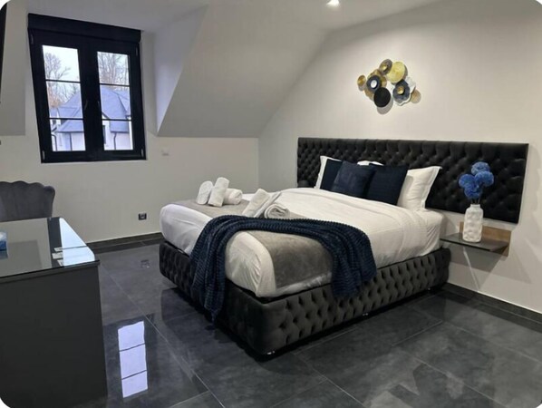 A modern bedroom with a tufted headboard, a large bed with white linens, a dark blue throw blanket, and a decorative wall piece above the bed. - Domaine de Bracheux (Fouquenies)