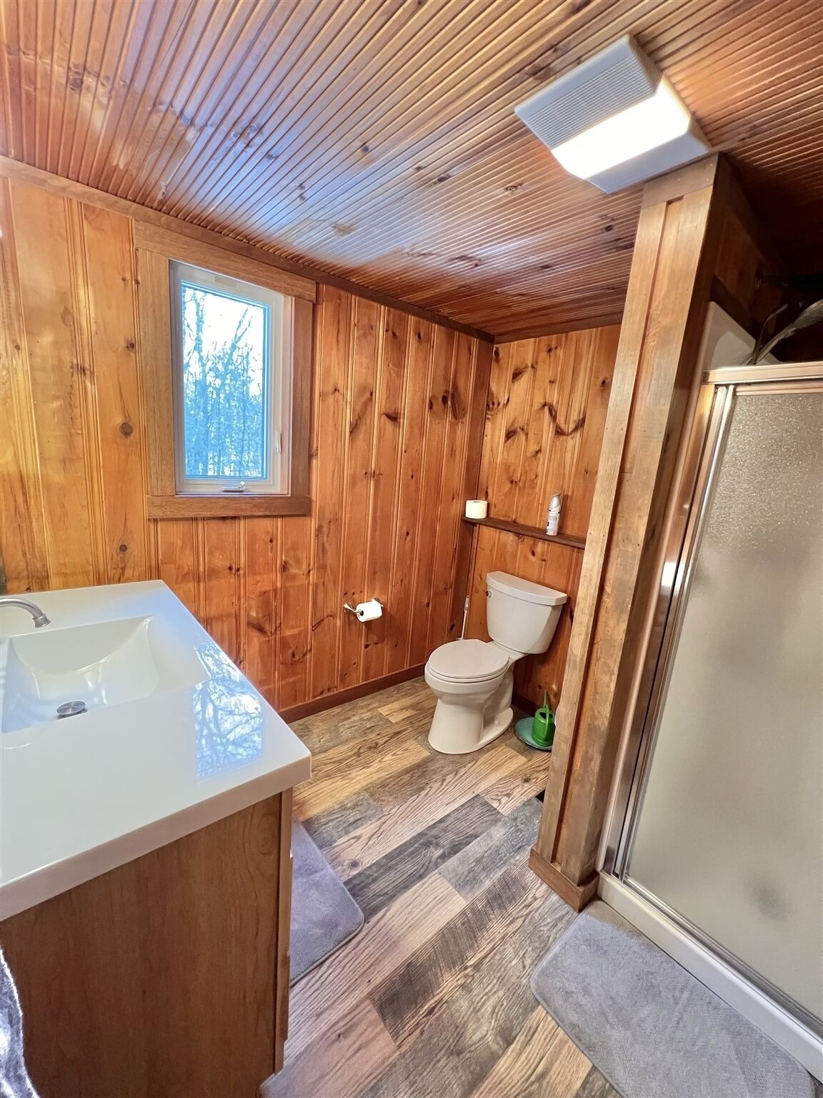 Une salle de bain aux murs lambrissés, avec un lavabo, des toilettes et une douche, le tout de couleur blanche.