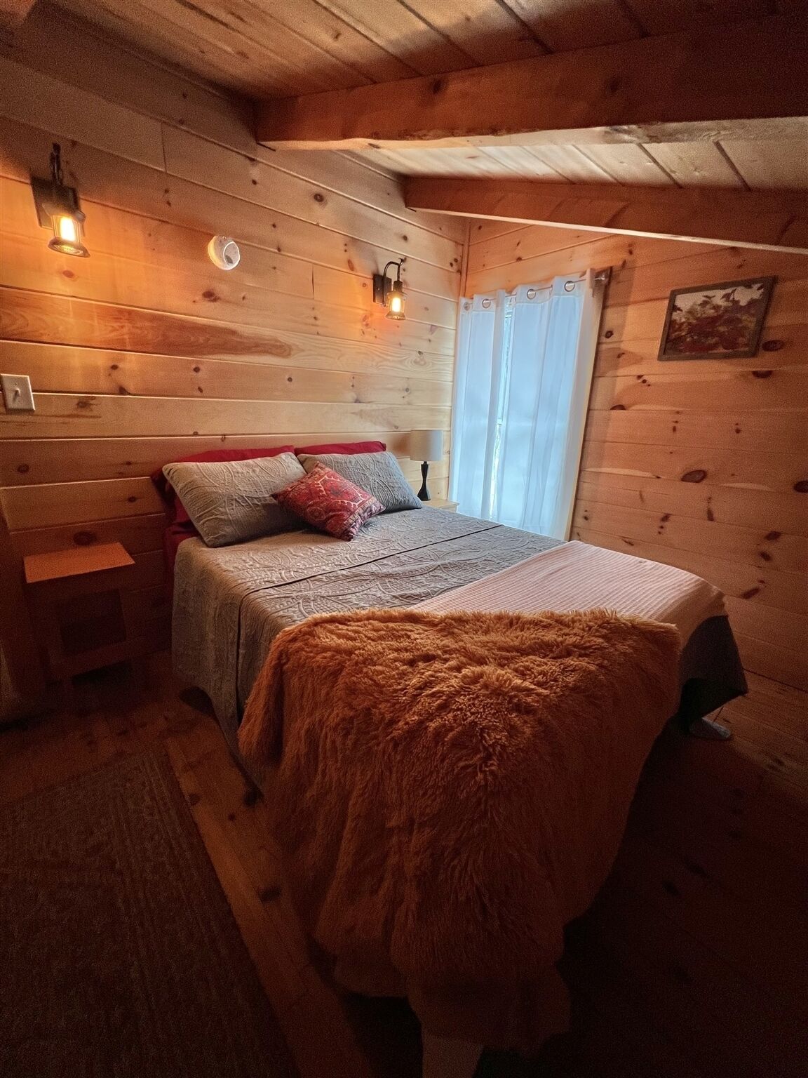 A cozy wooden cabin bedroom with a bed, a nightstand, a lamp, and a window with curtains.