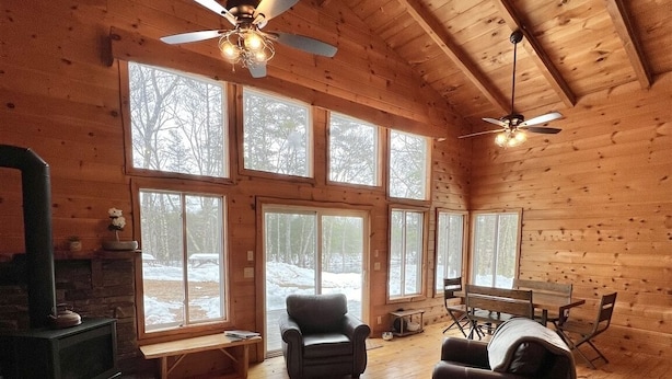 Interior acogedor de una cabaña con chimenea, sofá de cuero, mesa de comedor y ventanas amplias.