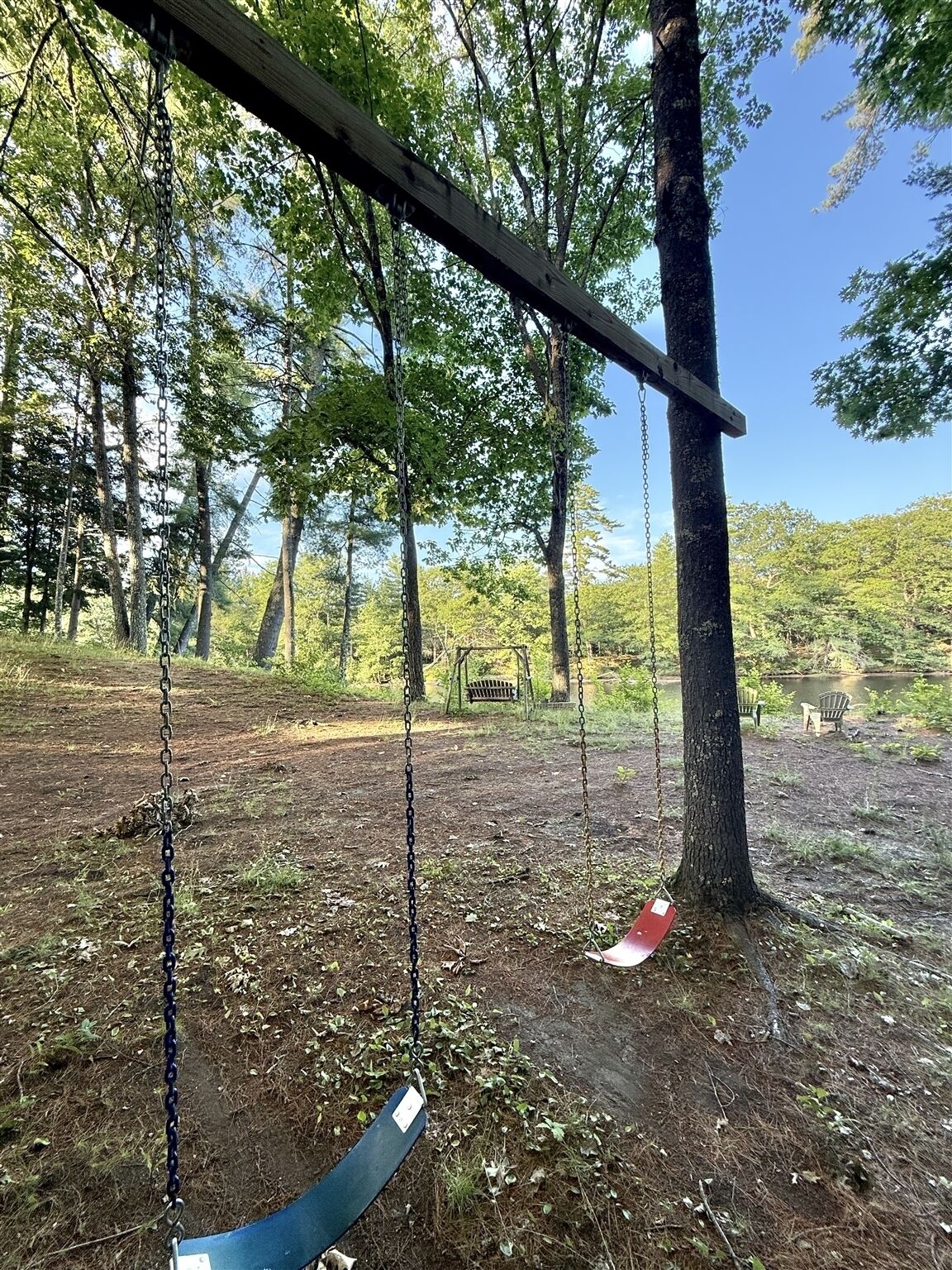 A swing set in a wooded area with a bench and chairs in the background.